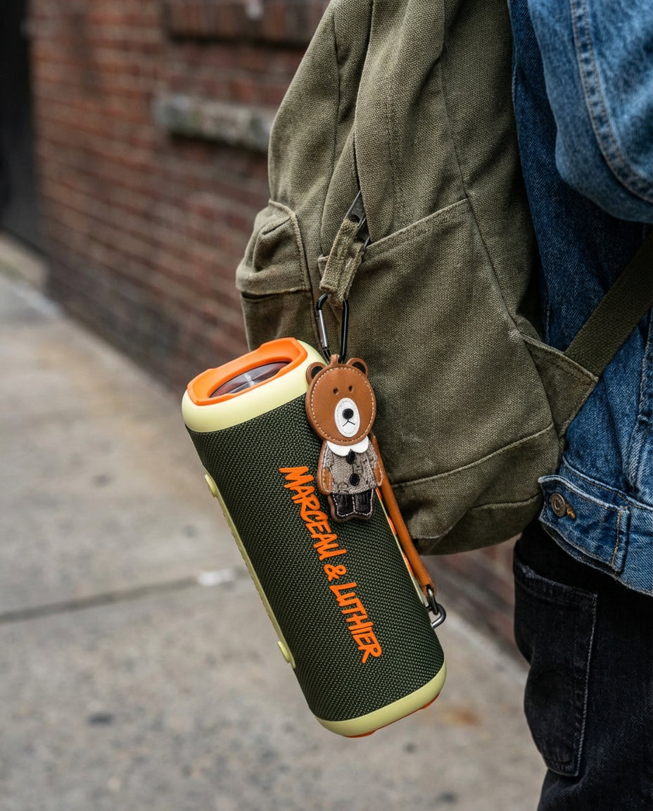 Altavoz portátil Nocturne de Marceau & Luthier colgado de una mochila de lona mediante un mosquetón, resaltando su portabilidad y el accesorio de oso decorativo.