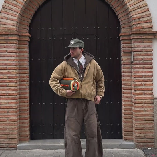 A video of a model standing in front of a large arched wooden door with brick detailing. He is wearing a tan corduroy jacket, a grey hoodie, a white shirt with a red tie, brown work pants, and a green cap. The model is holding the Nocturne portable speaker, which has an olive green body, orange branding, and a brown leather strap with a bear-shaped charm. The video shows him posing with the speaker under his arm and then walking away from the camera, carrying the speaker by its handle.