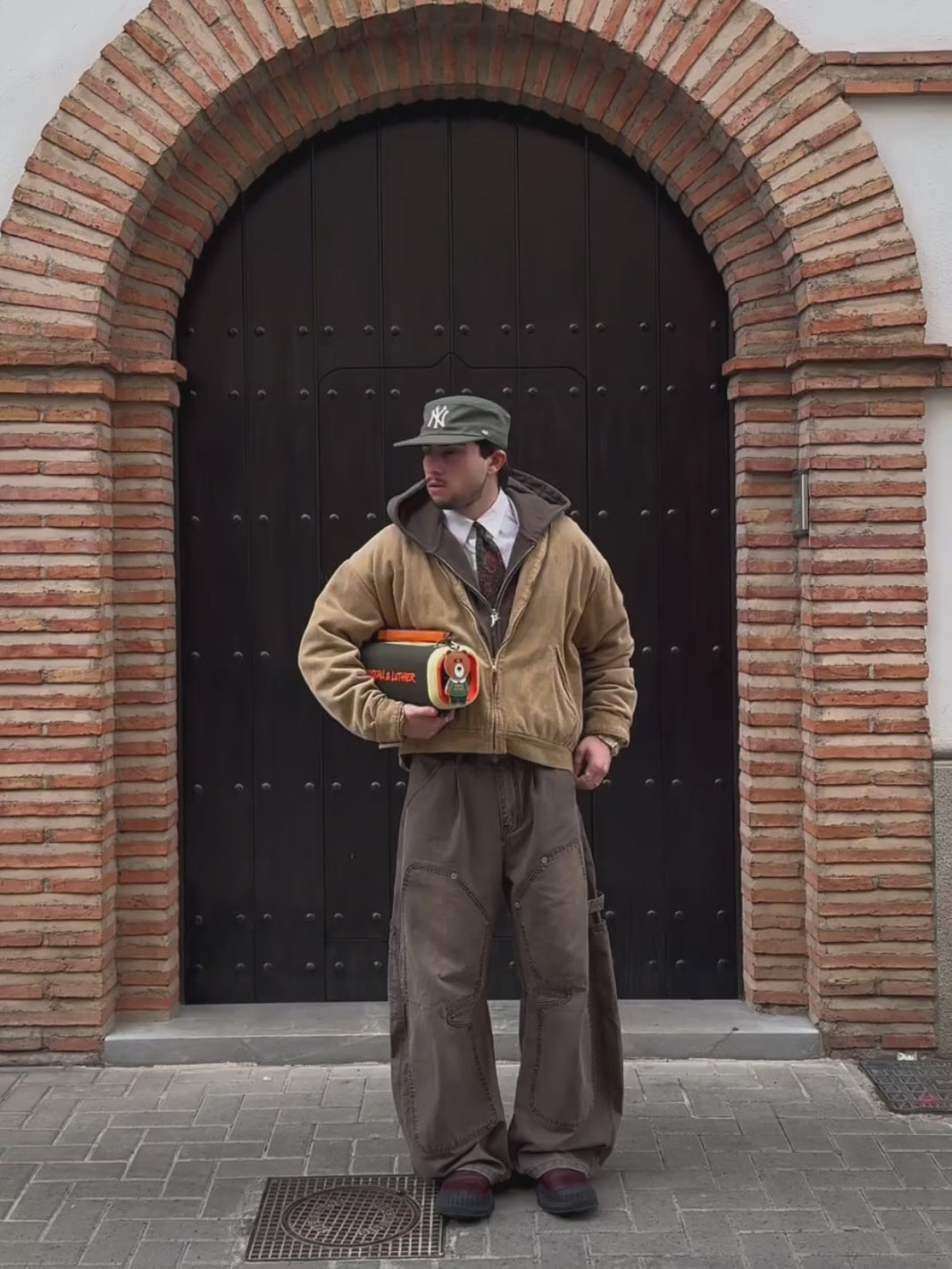 A video of a model standing in front of a large arched wooden door with brick detailing. He is wearing a tan corduroy jacket, a grey hoodie, a white shirt with a red tie, brown work pants, and a green cap. The model is holding the Nocturne portable speaker, which has an olive green body, orange branding, and a brown leather strap with a bear-shaped charm. The video shows him posing with the speaker under his arm and then walking away from the camera, carrying the speaker by its handle.
