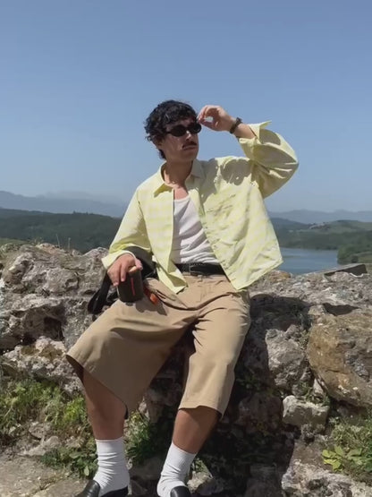 A man wearing a yellow plaid shirt and sunglasses sits on a rocky hill, holding a black and orange Marceau & Luthier BELLEVILLE portable bluetooth speaker. The video close-up shows the speaker's compact design and premium finish against a scenic landscape backdrop.