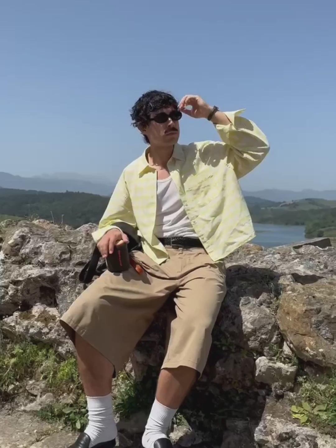 A man wearing a yellow plaid shirt and sunglasses sits on a rocky hill, holding a black and orange Marceau & Luthier BELLEVILLE portable bluetooth speaker. The video close-up shows the speaker's compact design and premium finish against a scenic landscape backdrop.