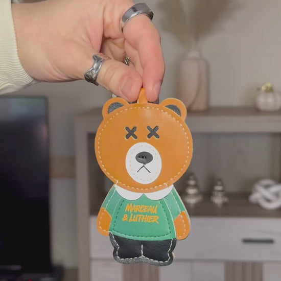 A video sequence showing a person's hands holding and rotating the PIGALLE portable speaker accessories. It starts with a close-up of a brown leather bear charm with "Marceau & Luthier" text, followed by an orange leather wrist strap. Finally, it shows the PIGALLE speaker, which features an olive green fabric body, orange embroidered branding, and a light yellow control panel with a power button and volume symbols. The background is a blurred living room with a shelf and decorative items.
