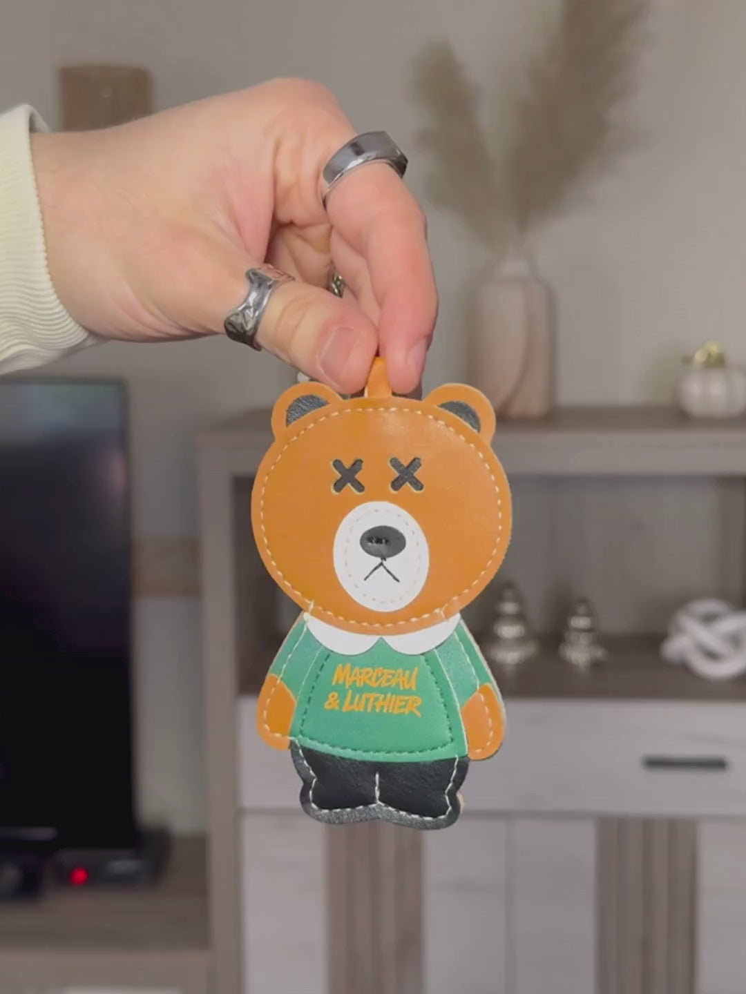 A video sequence showing a person's hands holding and rotating the PIGALLE portable speaker accessories. It starts with a close-up of a brown leather bear charm with "Marceau & Luthier" text, followed by an orange leather wrist strap. Finally, it shows the PIGALLE speaker, which features an olive green fabric body, orange embroidered branding, and a light yellow control panel with a power button and volume symbols. The background is a blurred living room with a shelf and decorative items.